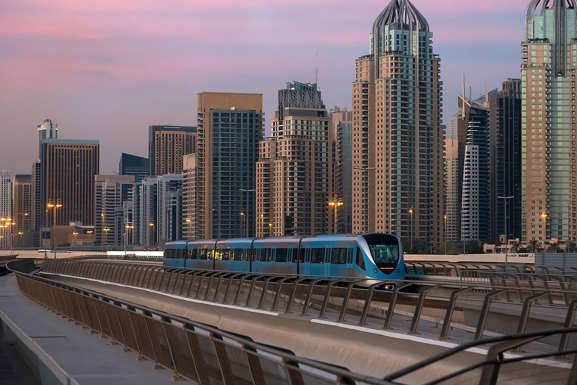 Un tren azul del Metro de Dubái deslizándose por su vía elevada al anochecer, con las torres de Dubai Marina detrás