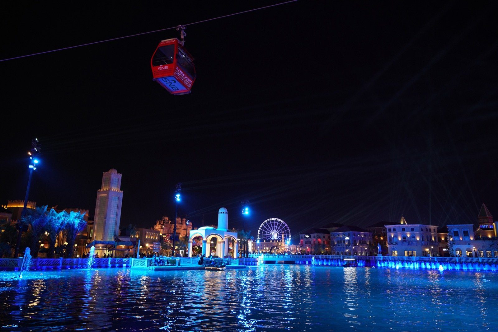 Boulevard Riyadh City iluminado de azul por la noche, con un teleférico que se desliza sobre su laguna y sus pabellones.
