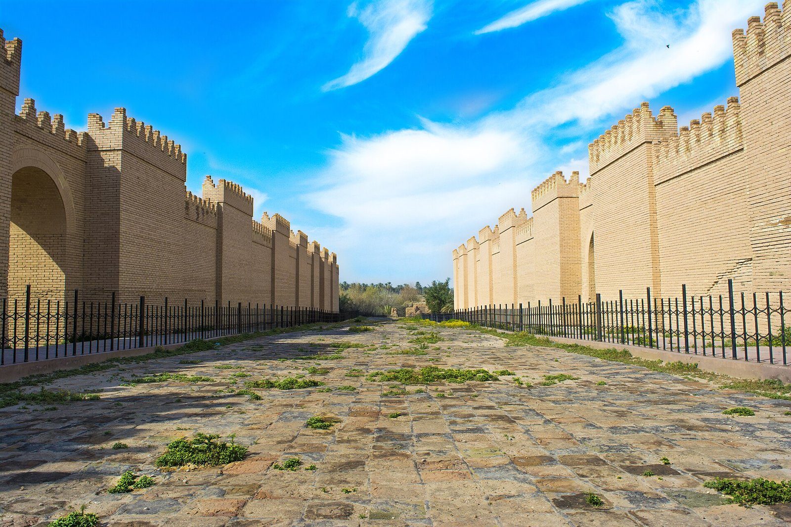 Las murallas almenadas reconstruidas de la antigua Babilonia, que flanquean una vía procesional pavimentada bajo un cielo de un azul intenso.