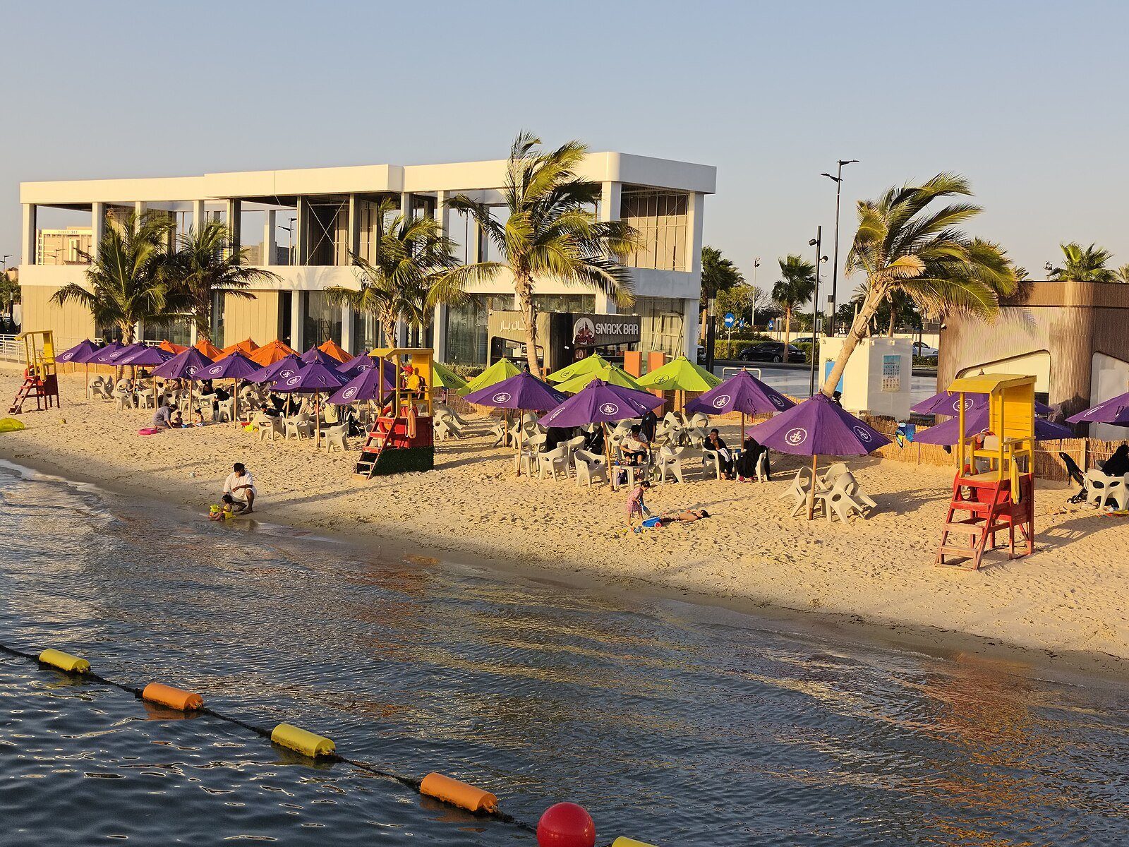 Un club de playa con sombrillas moradas y palmeras en el paseo marítimo de South Abhur, en Yeda, con la luz de última hora de la tarde.