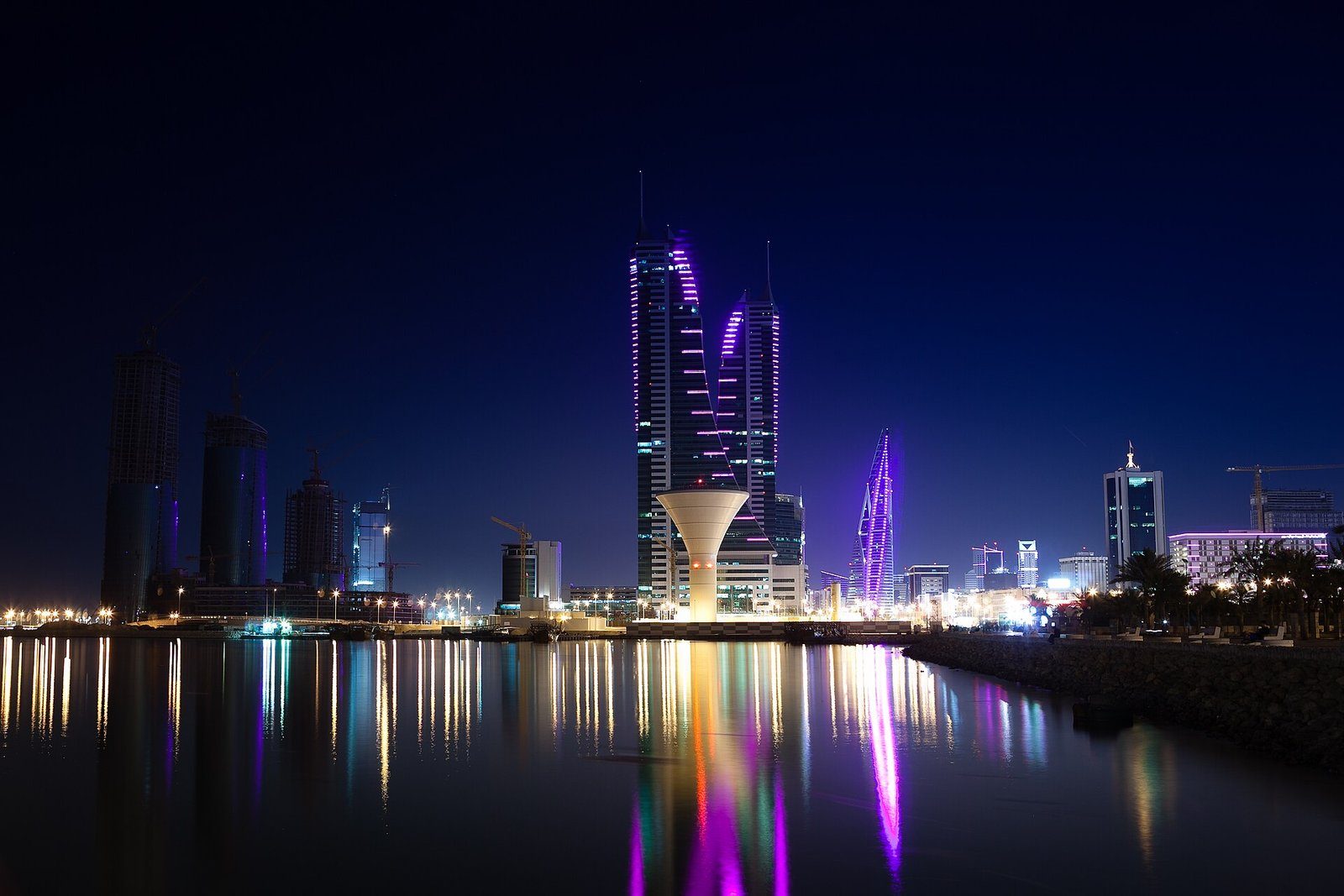 Las torres gemelas del Bahrain Financial Harbour iluminadas en violeta y reflejadas en las aguas tranquilas de Manama de noche.