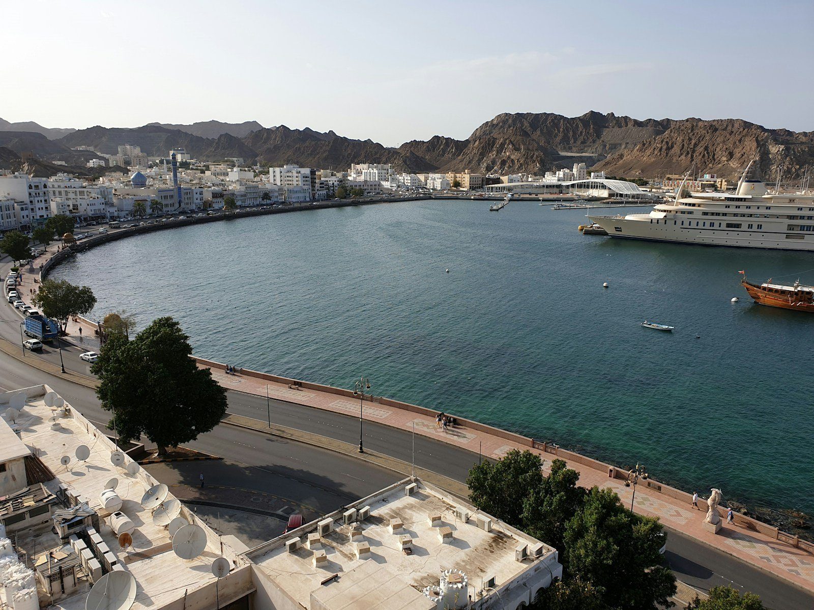 La curva de la Corniche de Muttrah y el puerto de Mascate, con un yate real y un dhow de madera al pie de las montañas de Hajar.