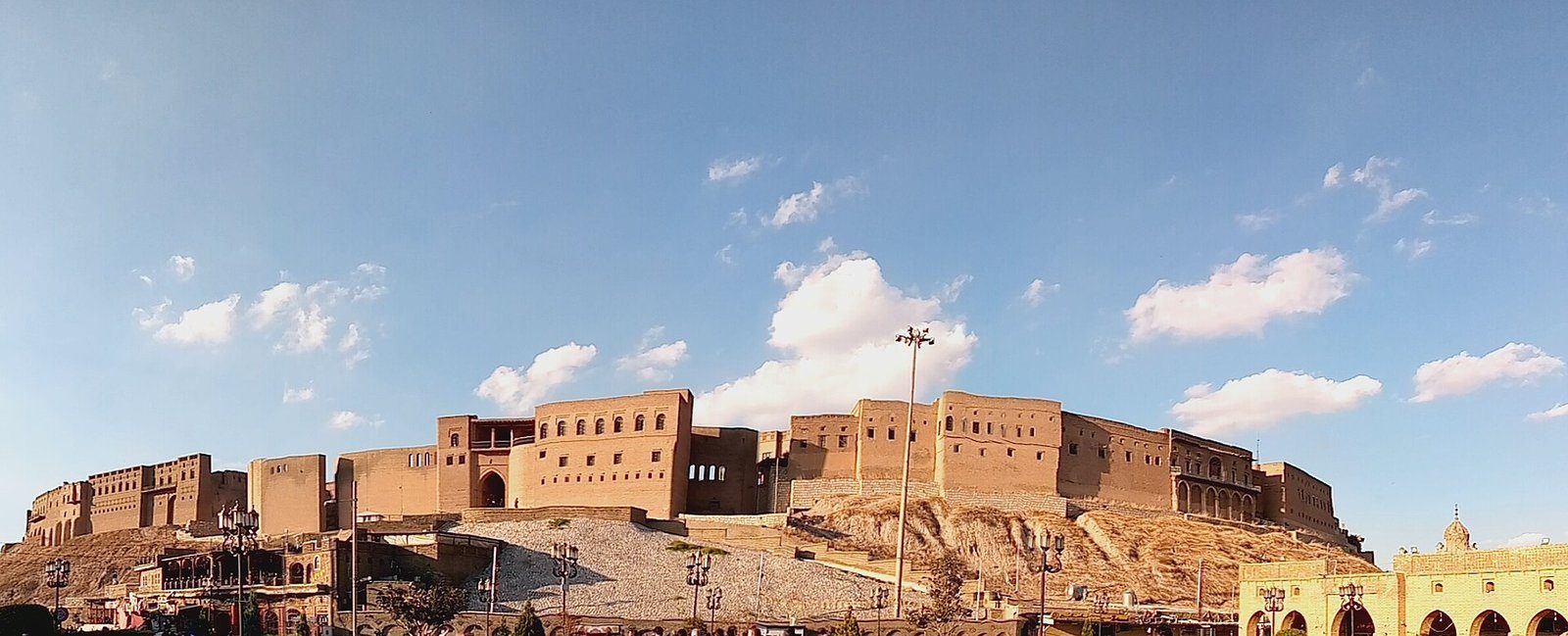 Las antiguas murallas de la Ciudadela de Erbil coronando su colina bajo un cielo azul con nubes blancas.