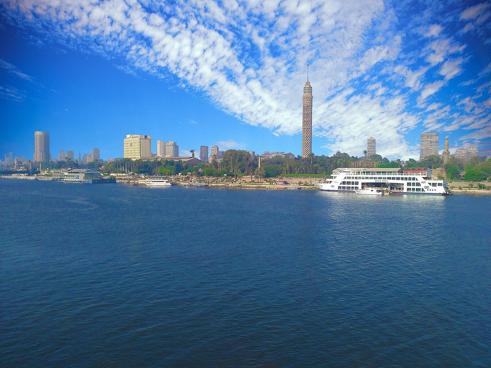 La Torre de El Cairo elevándose sobre la isla de Gezira y el Nilo azul profundo bajo un cielo de nubes blancas dispersas