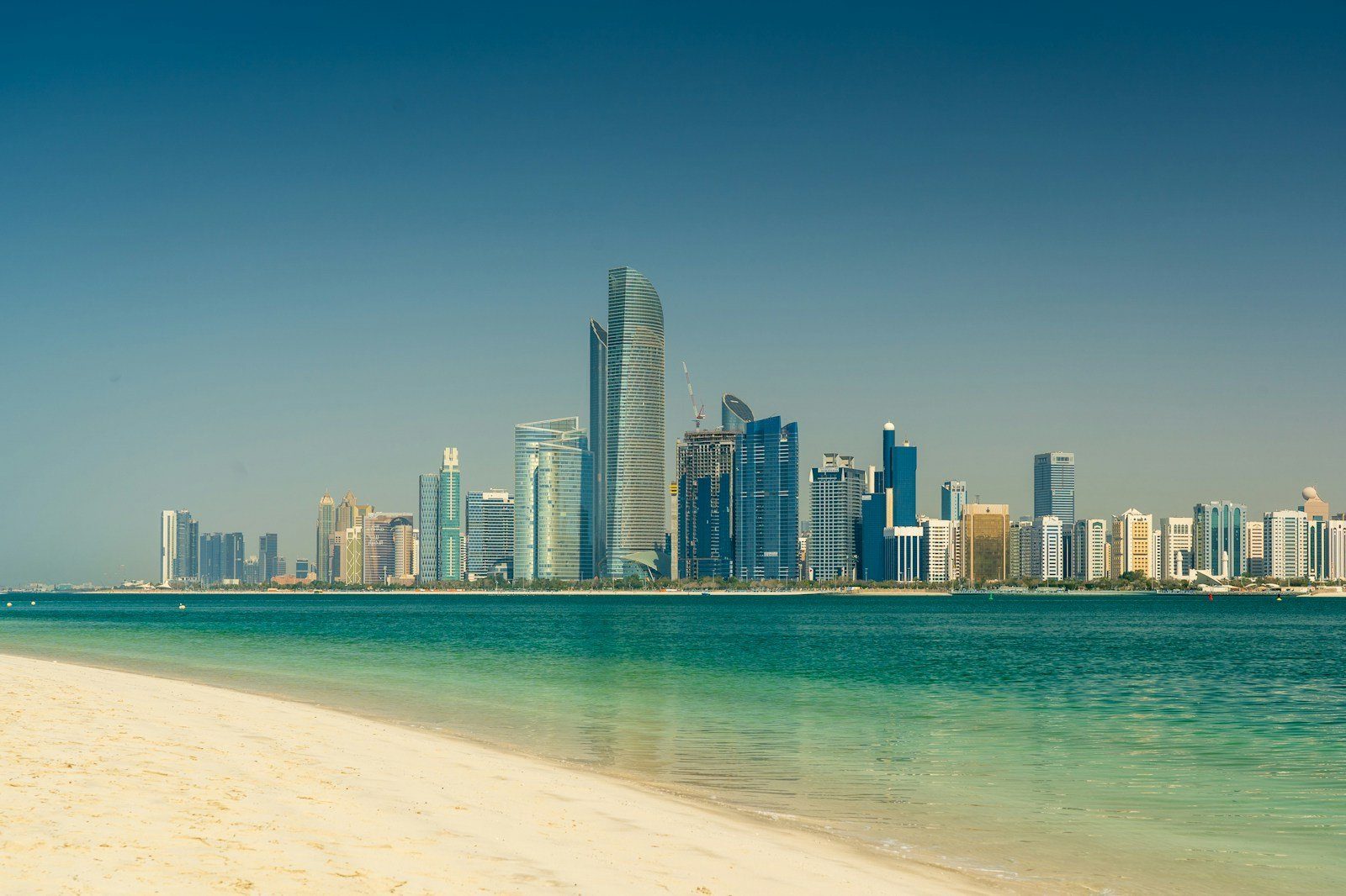 Las torres de la Corniche de Abu Dabi alzándose al otro lado de aguas turquesas y cristalinas desde una playa de arena blanca.