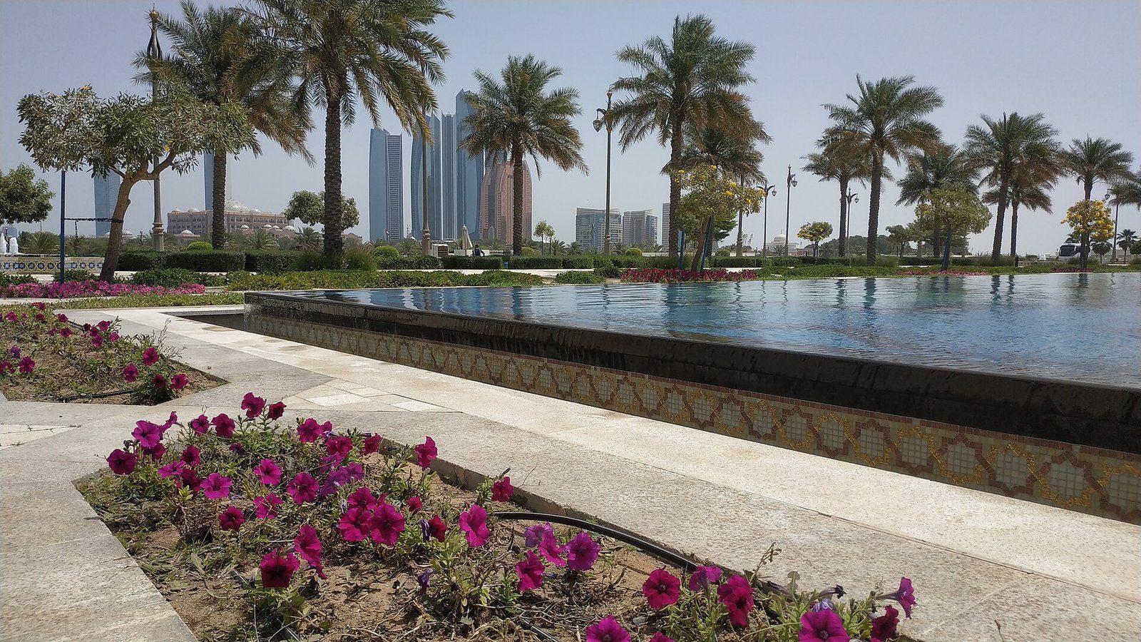 Un largo estanque reflectante, parterres de flores y palmeras datileras en los jardines de Qasr Al Watan, con las torres de Abu Dabi al fondo.