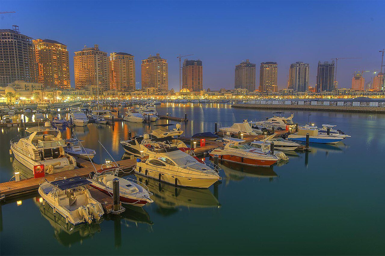 Yates amarrados en el puerto deportivo de The Pearl, en Doha, a la hora azul, con las torres del frente marítimo iluminadas detrás.