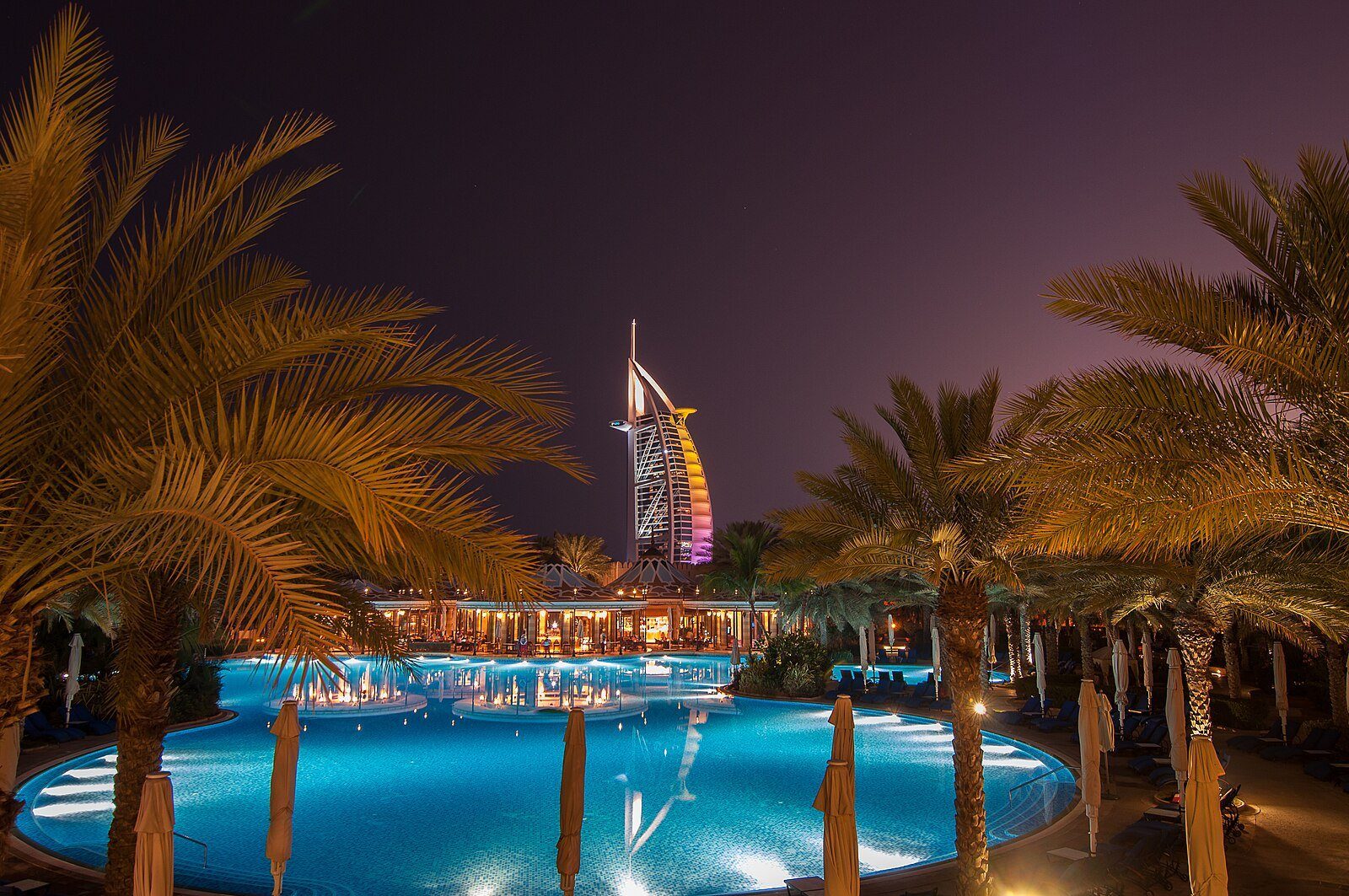 La vela iluminada del Burj Al Arab alzándose más allá de las palmeras y de una piscina de resort suavemente iluminada al anochecer en Dubái