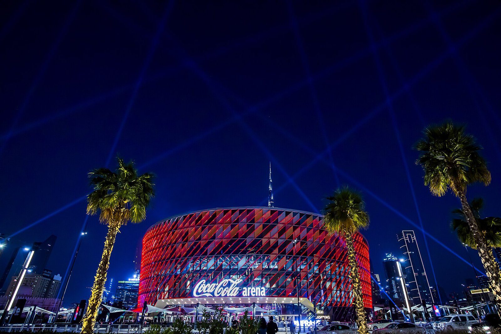 El Coca-Cola Arena de Dubái iluminado en rojo de noche, con reflectores y el Burj Khalifa más allá de las palmeras