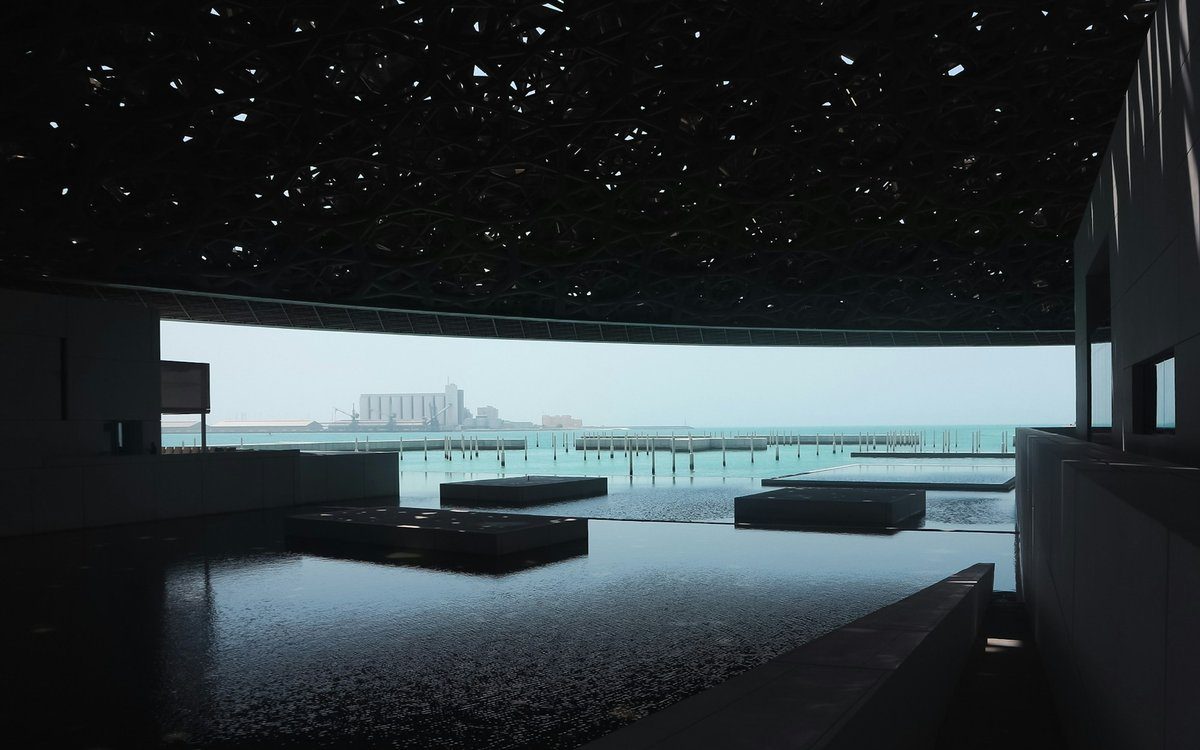 La luz cae a través de la cúpula reticulada del Louvre Abu Dhabi sobre aguas quietas que se abren al Golfo.