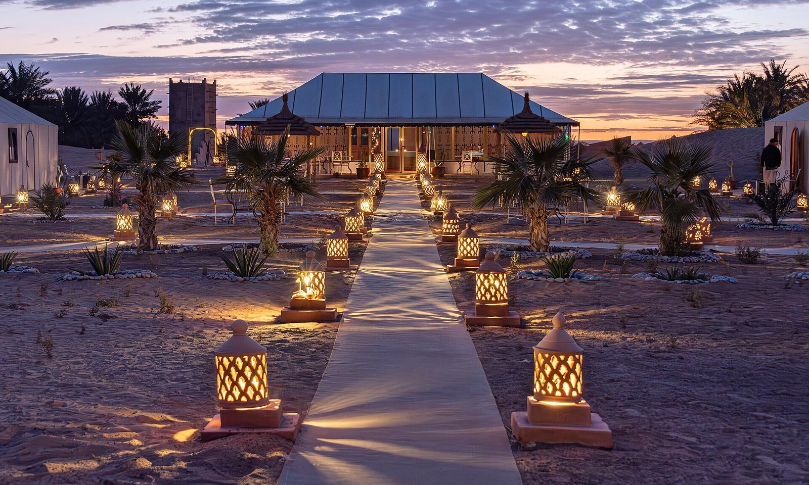 Senderos iluminados con faroles y palmeras en un campamento de lujo bajo las dunas de Erg Chebbi al anochecer, Merzouga