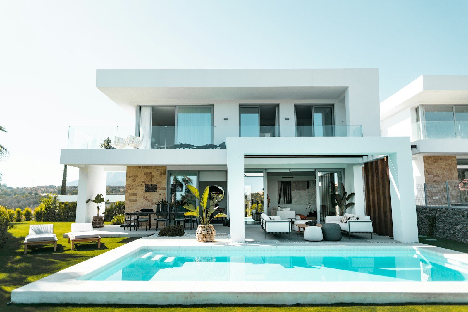 Una villa blanca moderna con un salón al aire libre y tumbonas junto a su piscina privada y su césped.