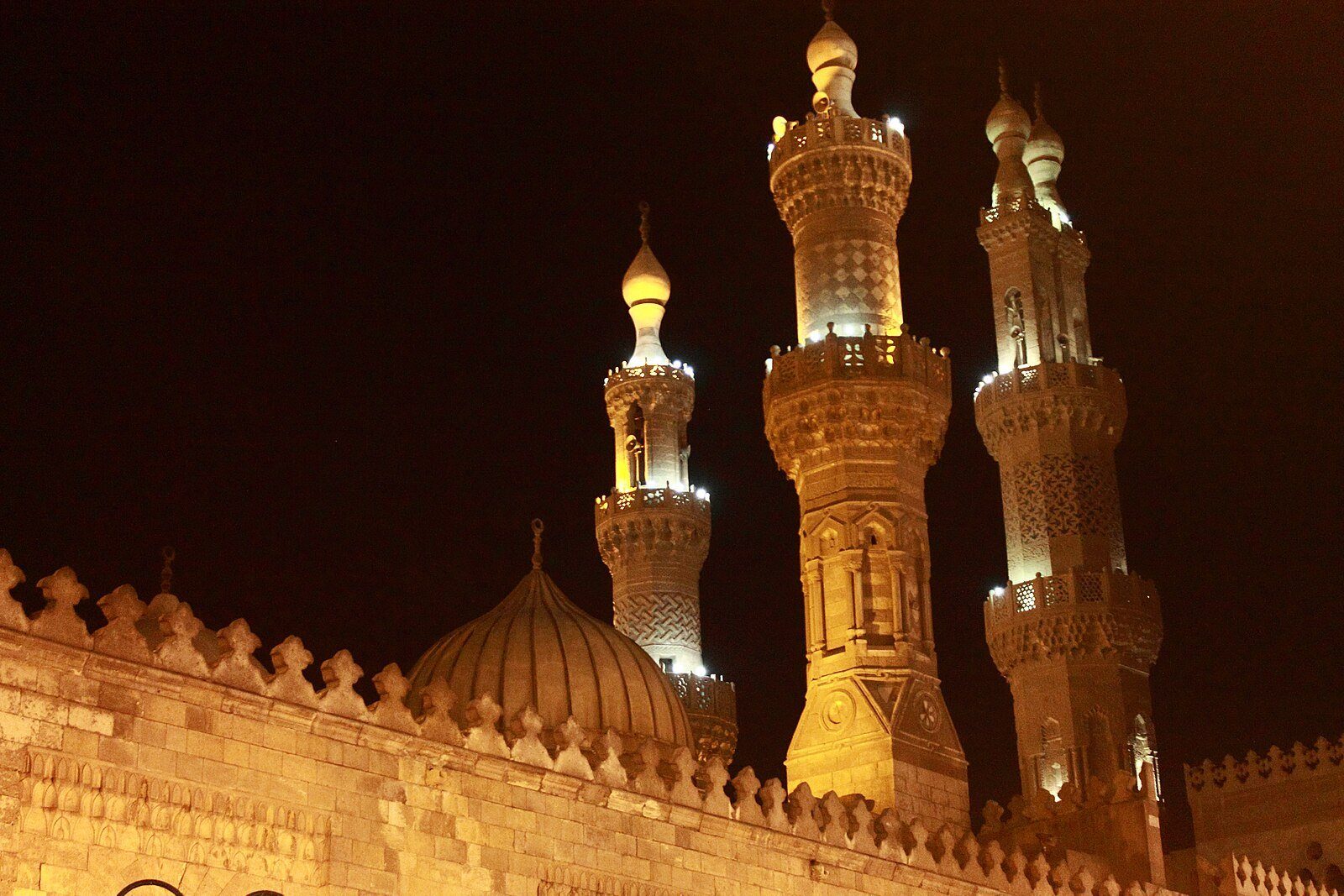 Minaretes mamelucos iluminados y una cúpula de piedra acanalada sobre los muros almenados de una mezquita en El Cairo islámico de noche