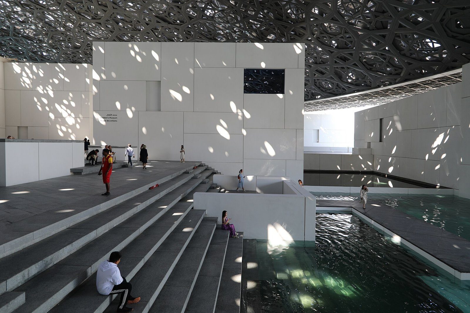 La luz del sol se filtra por la cúpula calada sobre las galerías blancas y el agua del Louvre Abu Dhabi